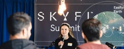 Payton Barnwell speaking with a microphone in front of a SkyFi Summit banner.